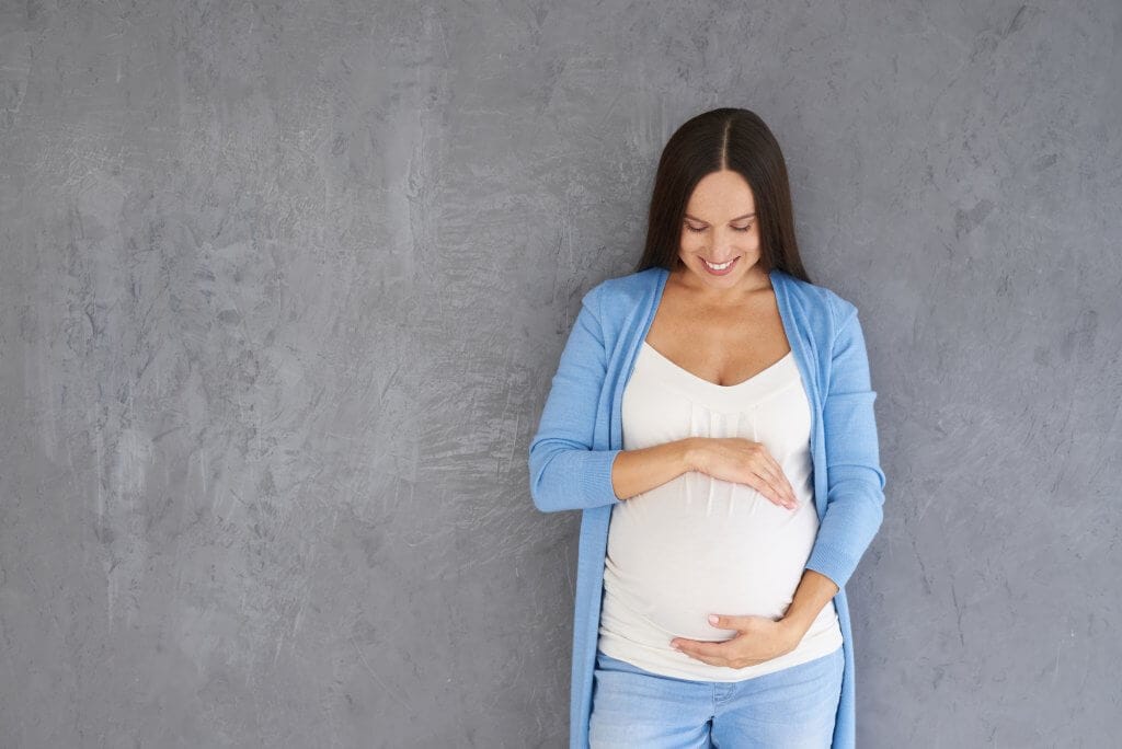 Pregnant woman cradling belly, smiling in blue sweater.
