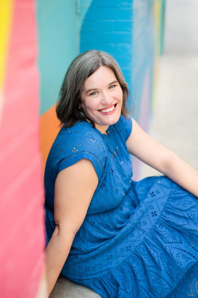 Carmen smiling in blue dress against colorful wall.